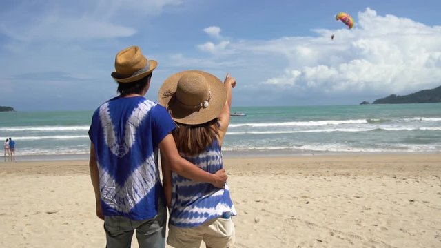 Young couple traveler looking parachute flying in the sky on the beach, Couple vacation travel summer concept