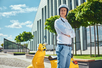 Confident handsome man with stylish haircut in a white shirt, jeans, and motocycle helmet standing with crossed arms near a yellow classic italian scooter against a skyscraper.