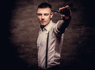 Portrait of a old-fashioned tattooed hairdresser wearing a white shirt with suspenders holds a scissors. Isolated on dark background.