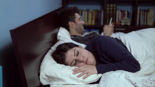 irritated woman in bed scolding husband playing loudly with smartphone 