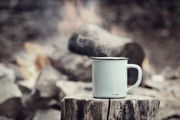 Cup of hot steaming coffee sitting on an old log by an outdoor campfire with a vintage antique...