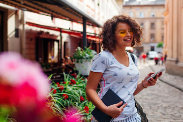 Beautiful college girl using smart phone and holding book outdoors