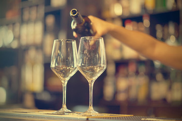 A barman girl standing smiling, pours white wine into a glass from a bottle. Shelves with bottles of alcohol in the background. Focus on the glass