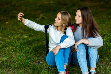 Fototapeta premium Two teenage girls. In the summer of the city park. They sit on the grass and take photos on smartphone. Behind backpacks. The concept of school friendship. Emotion pose on the phone.