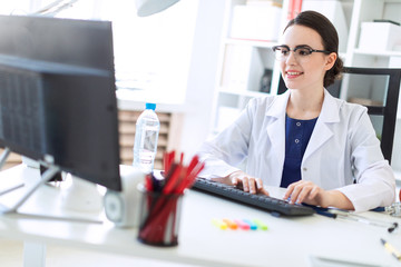 A beautiful young girl in a white robe is sitting at the table and typing on the keyboard.