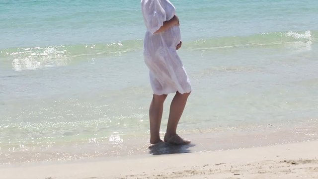 Young pregnant woman in white dress walking on the beach near blue sea and breathing. Summer vacation during pregnancy, happy motherhood concept