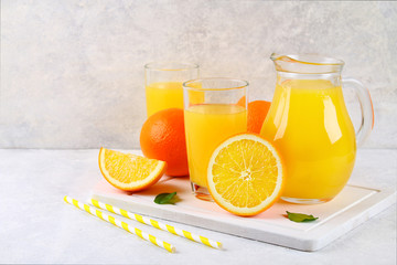 Glass cups and a pitcher of fresh orange juice with slices of orange and yellow tubes on a light gray table.