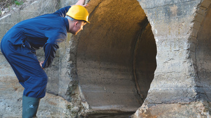 Engineer, worker checks gutters, in blue robe, in boots, in a construction yellow helmet, tunnels, technical supervision.