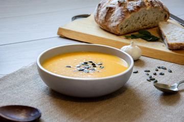 Pumpkin soup with pumpkin seeds. In white bowl, on hessian fabric. With wooden spoon, garlic and wholegrain rustic bread on wooden board. 