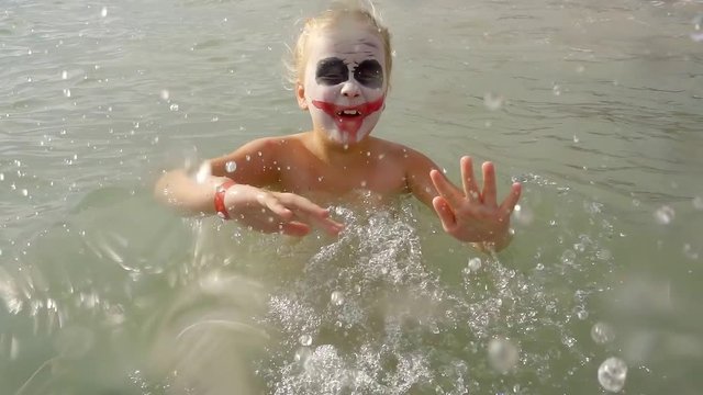 little girl with face art joker style is bathing in sea water and splashing in camera, shouting and grimacing