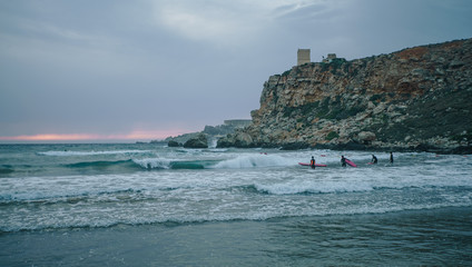 MALTA - June 15, 2018: Surfers with the surfboards catching big waves in the evening in the sea. Beautiful sunset on the beach.