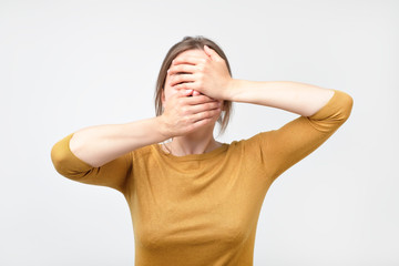 Young european woman in yellow sweater hides her face, studio photo isolated on gray background. She has social phobia, tying to be anonym