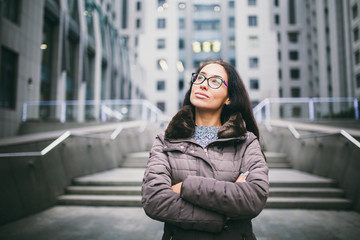 Beautiful young woman of European ethnicity with long brunette hair looking to side and up, wearing glasses and coat stands on background of the business center in the fall in cloudy cloudy weather