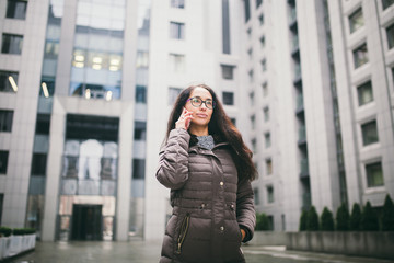 Fototapeta premium Theme is the business situation. Beautiful young woman of European ethnicity with long brunette hair wearing glasses and coat stands on background of business center and uses phone in hand near ear
