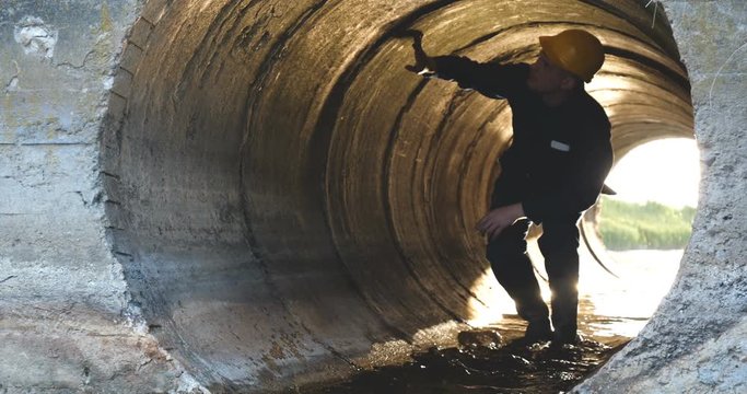 Engineer, worker checks gutters, in blue robe, in boots, in a construction yellow helmet, tunnels, technical supervision.