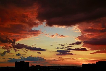 Dark colorful dramatic sky with purple and pink clouds, red and golden sunset behind houses, black...