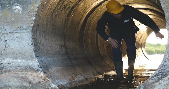 Engineer, worker checks gutters, in blue robe, in boots, in a construction yellow helmet, tunnels, technical supervision.