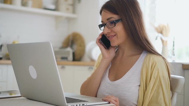 Young female self-employed sales manager working on brand-new laptop and having phone talk in tha tkitche. Indoors. Portrait.