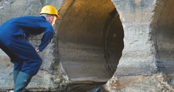 Engineer, worker checks gutters, in blue robe, in boots, in a construction yellow helmet, tunnels, technical supervision.