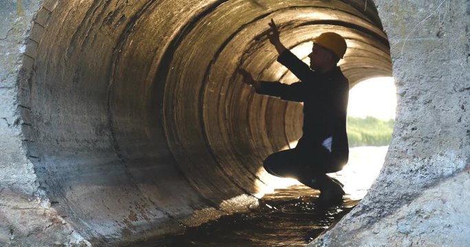 Engineer, worker checks gutters, in blue robe, in boots, in a construction yellow helmet, tunnels, technical supervision.
