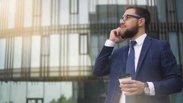 Successful business owner talking on phone, drinking coffee standing on city street, bearded businessman is having business dialogue, enjoying hot drink on background of modern building. Concept
