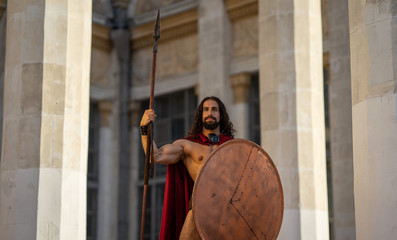 a Spartan warrior with a spear and shield. buildings with columns on the background.