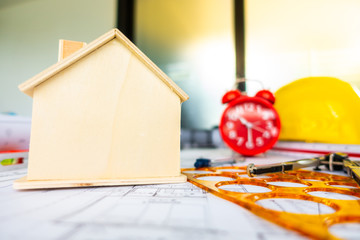 Red clock on archetecture blueprint with pencil, yellow helmet, wooden house model and engineering equipments in modern working office room