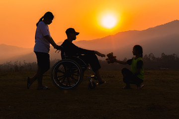 Fototapeta na wymiar silhouetts disabled man is sitting on wheelchairs at sunset time with children helping he.