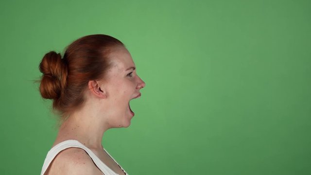 Profile portrait of a young red haired woman screaming furiously on green chromakey background. Ginger haired female shouting angrily. Stress, depression, nervous breakdown concept.