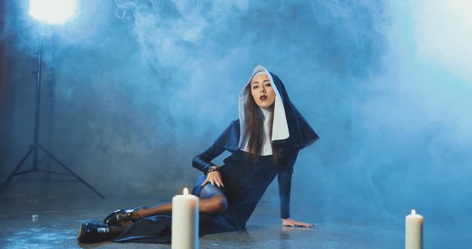 Sexual nun is seductively sitting on the floor and stroking her leg in stockings in a dark smoke studio among the candles