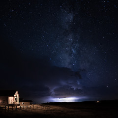 Naklejka premium A wood cabin with the porch light on with a wooden fence and grass field with a lightning storm and dramatic clouds behind it with the Milky Way galaxy rising above it in a starry night sky