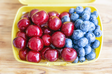 Ripe fresh tasty bio cherries and bluberries in plastic lunchbox . Organic sweet berries takeaway quick food for picnic. Healthy snacks. Wooden table on background