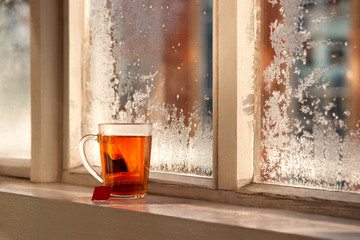 Tea on the winter window sill. Tea and tea bag against the background of frost on the window. A transparent mug of tea on the balcony window sill in a cold winter morning.