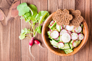 Obraz na płótnie Canvas Lenten spring vegetable salad of cucumber, radish, greens and rye bread in a plate on a wooden table. Top view