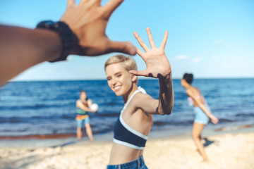 partial view of smiling woman giving high five to man while multicultural friends playing volleyball on beach