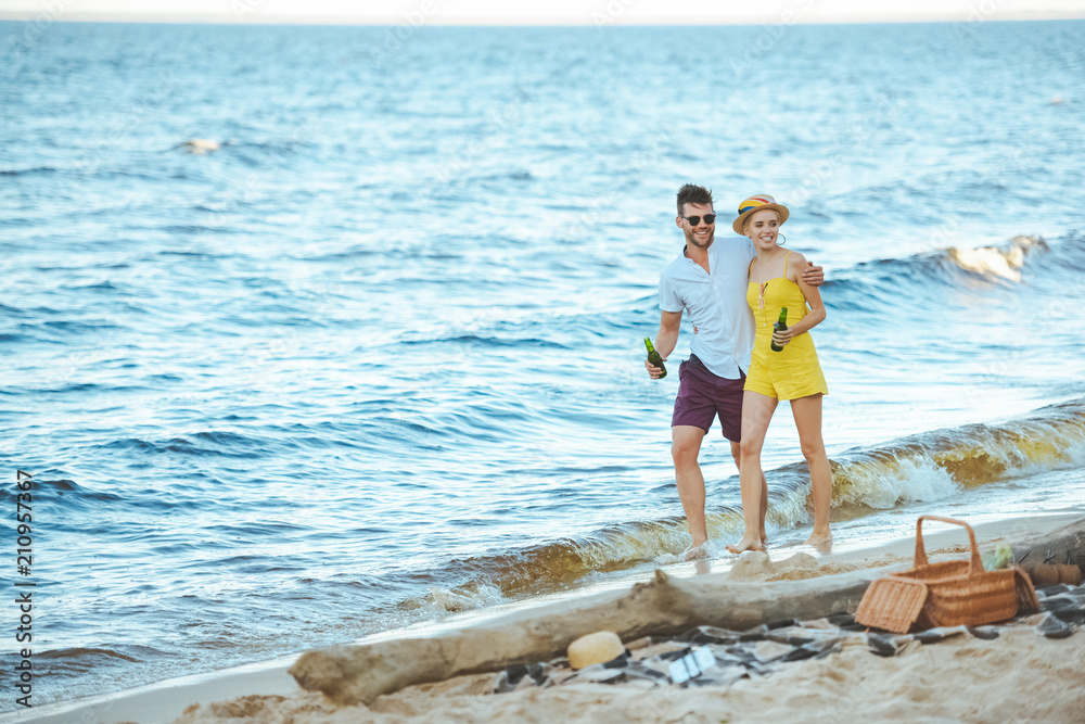 Wall mural cheerful couple with beer in glass bottles walking on beach