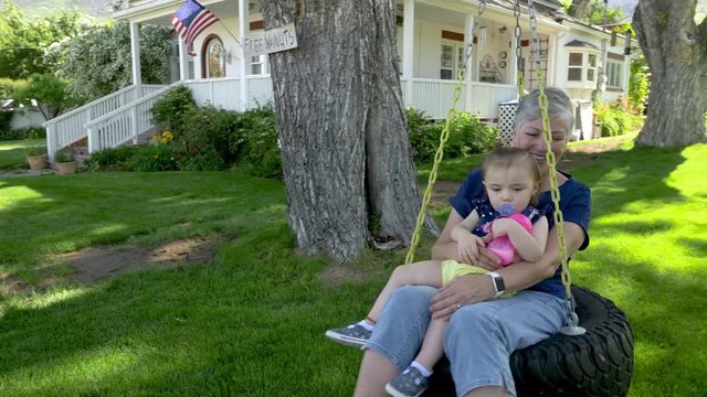 Grandma holding toddler in tire swing in yard in the shade on sunny day.