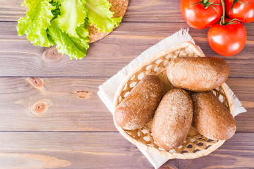 Fresh rye buns in a basket, salad and tomato on a wooden table. Top view. Copy space