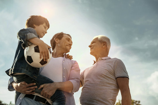 Feeling proud. Exuberant smiling loving daddy son and grandfather looking at each other while the dad holing his son in his hands and the son holding his ball