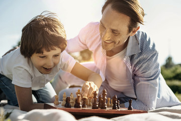 Favourite game. Joyful laughing kneeling boy playing chess with his dad enjoying the nature and his dad smiling