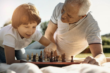 Making a move. Joyful concentrated young boy playing chess with his grandpa and making a move while his grandpa watching him