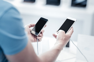 Comparison. Attentive responsible careful customer comparing two modern smartphones in the electronics shop
