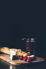 front view of brie, raspberries, jam in jar and baguette on cutting board on black
