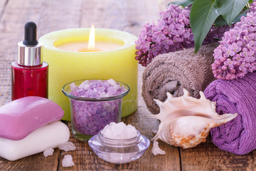 Soap, red bottle with aromatic oil, burning candle, bowls with sea salt,  sea shell, lilac flowers and towels