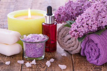 Obraz na płótnie Canvas Soap, burning candle, bowl with sea salt, red bottle with aromatic oil, lilac flowers and towels on wooden boards