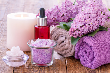 Bowls with sea salt, burning candle, red bottle with aromatic oil, handmade soap, lilac flowers and towels for bathroom procedures
