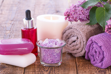 Obraz na płótnie Canvas Red bottle with aromatic oil, burning candle, soap, bowl with sea salt, lilac flowers and towels