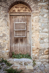 Fototapeta na wymiar Old brown wooden door and dirty stone wall. Plants growing and two stone steps. Entrance to the house 