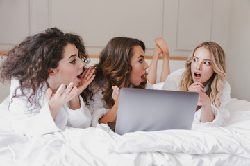 Image of excited surprised women 20s wearing white housecoat lying in luxury bedroom at home, and using silver laptop during hen party