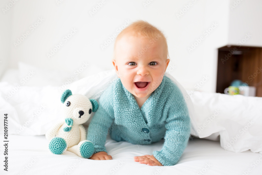 Wall mural Little cute baby boy, child in knitted sweater, holding knitted toy, smiling happily at camera